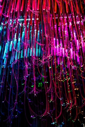 Colorful strands of fiber optic cables cascade downwards, illuminated in vibrant shades of pink, blue, and green. The strands are interspersed with small sparkling lights, creating a dazzling display of light and color.