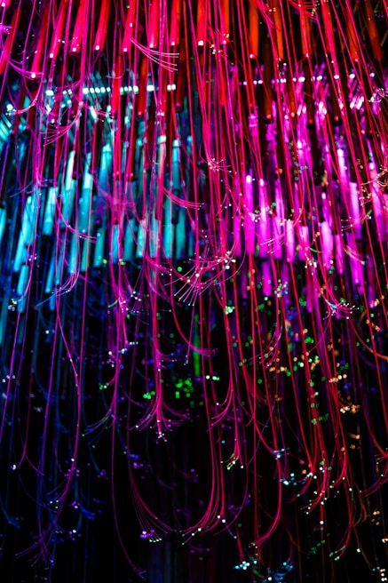Colorful strands of fiber optic cables cascade downwards, illuminated in vibrant shades of pink, blue, and green. The strands are interspersed with small sparkling lights, creating a dazzling display of light and color.