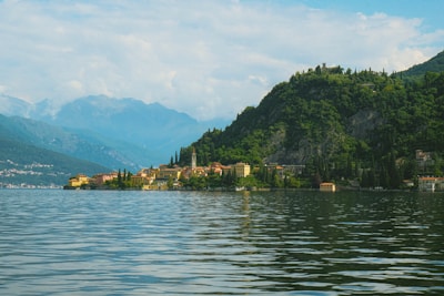 A picturesque lakeside view with a serene body of water in the foreground. Along the shore, a quaint village with colorful buildings nestles into the landscape, while lush green hills rise behind them. The backdrop features distant, hazy blue mountains under a partly cloudy sky.