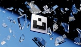 Floating geometric shapes, resembling shattered glass, are suspended around a central logo against a backdrop transitioning from black to blue. The scene conveys a sense of motion and fragmentation.