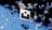 Floating geometric shapes, resembling shattered glass, are suspended around a central logo against a backdrop transitioning from black to blue. The scene conveys a sense of motion and fragmentation.