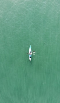 A lone person is kayaking on calm, green water. The kayak is white and blue, and the individual is seen from an overhead perspective. The water beneath has a subtle texture.