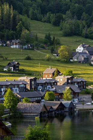 A serene village with rustic, wooden houses situated near a calm body of water. The landscape includes green hills and dense forest in the background, highlighted by sunlight casting soft shadows. The architecture reflects traditional design, nestled harmoniously within lush greenery.