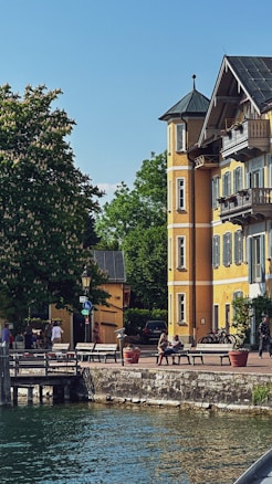 A picturesque lakeside scene featuring a charming, yellow building with a turret and traditional architecture. Several people are sitting and walking near the water, enjoying the sunny day. Lush green trees and a clear blue sky add to the tranquil atmosphere. Planters and benches line the waterfront, and bicycles are parked nearby.