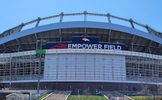 The image features a large sports stadium with a prominent sign that reads 'Empower Field'. The structure is primarily composed of metal and glass elements. A set of numbered signs for parking or entry is visible, along with steps leading up to the venue. The sky is clear, indicating a sunny day.