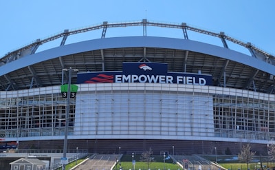The image features a large sports stadium with a prominent sign that reads 'Empower Field'. The structure is primarily composed of metal and glass elements. A set of numbered signs for parking or entry is visible, along with steps leading up to the venue. The sky is clear, indicating a sunny day.