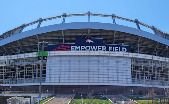 The image features a large sports stadium with a prominent sign that reads 'Empower Field'. The structure is primarily composed of metal and glass elements. A set of numbered signs for parking or entry is visible, along with steps leading up to the venue. The sky is clear, indicating a sunny day.