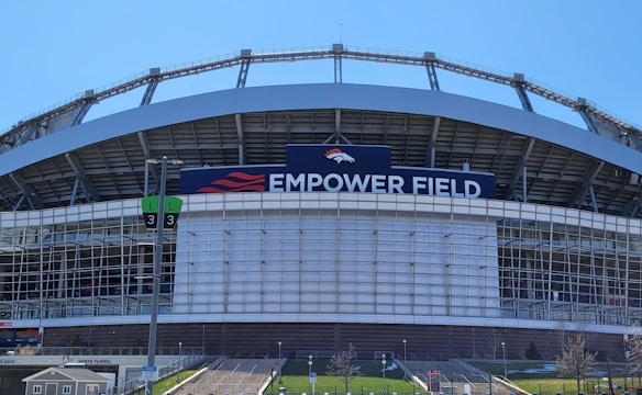 The image features a large sports stadium with a prominent sign that reads 'Empower Field'. The structure is primarily composed of metal and glass elements. A set of numbered signs for parking or entry is visible, along with steps leading up to the venue. The sky is clear, indicating a sunny day.