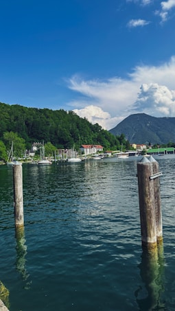 The image features a serene lake with clear blue water, surrounded by lush green trees and mountains in the background. A few boats can be seen docked near a quaint village on the shore. Two wooden posts are in the foreground, partially submerged in water under a bright blue sky with scattered clouds.