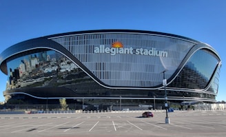 A modern, sleek stadium with a curved, futuristic design featuring large reflective glass surfaces. The name 'allegiant stadium' is prominently displayed on the structure. The stadium is situated in an expansive area with a largely empty parking lot in front. The clear blue sky above enhances the reflective quality of the building's facade.