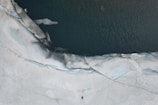 An aerial view of an ice-covered landscape with a body of dark water cutting through. The ice appears to be cracked and melting at the edges where it meets the water.