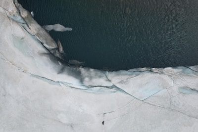 An aerial view of an ice-covered landscape with a body of dark water cutting through. The ice appears to be cracked and melting at the edges where it meets the water.