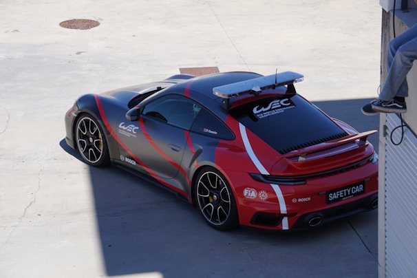 A sports car with a sleek, aerodynamic design is parked on a concrete surface. The car features a red and black color scheme with white racing stripes and logos, including 'WEC' and FIA markings, indicating its role as a safety car. A sophisticated light bar is mounted on the roof. A person is visible sitting on a ledge in the top right corner, casually wearing jeans and sneakers.