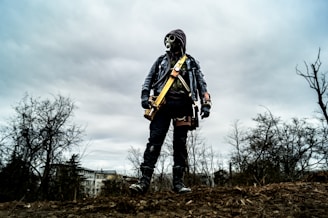 A person wearing a mask and gloves collecting soil samples outdoors with chemtrail clouds overhead