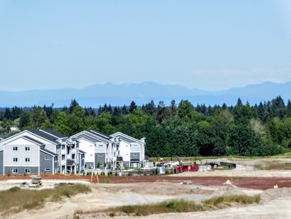 A newly constructed residential complex with multiple identical modern buildings sits in front of a lush forest. The scene is set against a clear blue sky and distant mountains, with a construction site visible in the foreground and several shipping containers and construction materials scattered around.