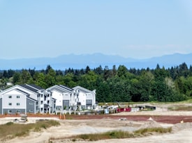 A newly constructed residential complex with multiple identical modern buildings sits in front of a lush forest. The scene is set against a clear blue sky and distant mountains, with a construction site visible in the foreground and several shipping containers and construction materials scattered around.