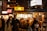 A bustling outdoor food market at night, with people gathered around a stall illuminated by warm lighting. Many individuals are wearing face masks and there are neon signs and traditional lanterns in the background, adding to the vibrant atmosphere.