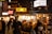 A bustling outdoor food market at night, with people gathered around a stall illuminated by warm lighting. Many individuals are wearing face masks and there are neon signs and traditional lanterns in the background, adding to the vibrant atmosphere.