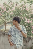 A person stands outdoors wearing a tropical print shirt and looking to the side. Pink flowers bloom on the trees in the background, creating a serene and natural setting.