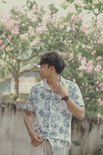A person stands outdoors wearing a tropical print shirt and looking to the side. Pink flowers bloom on the trees in the background, creating a serene and natural setting.