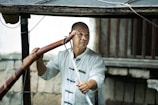 A person wearing a traditional martial arts uniform holds a wooden staff. The background includes wooden beams and a stone wall, suggesting an outdoor setting or rustic environment.
