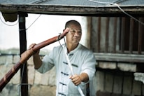 A person wearing a traditional martial arts uniform holds a wooden staff. The background includes wooden beams and a stone wall, suggesting an outdoor setting or rustic environment.