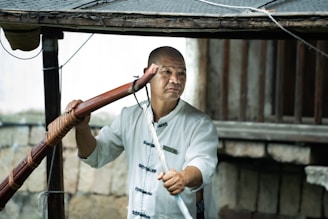A person wearing a traditional martial arts uniform holds a wooden staff. The background includes wooden beams and a stone wall, suggesting an outdoor setting or rustic environment.