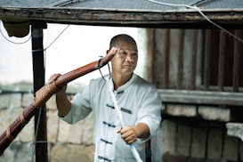 A person wearing a traditional martial arts uniform holds a wooden staff. The background includes wooden beams and a stone wall, suggesting an outdoor setting or rustic environment.