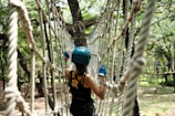 A person wearing a blue helmet and gloves is engaged in a rope course in a wooded area. The person appears to be carefully navigating through a suspended network of ropes, surrounded by trees with sunlight filtering through the leaves.