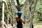 A person wearing a blue helmet and gloves is engaged in a rope course in a wooded area. The person appears to be carefully navigating through a suspended network of ropes, surrounded by trees with sunlight filtering through the leaves.