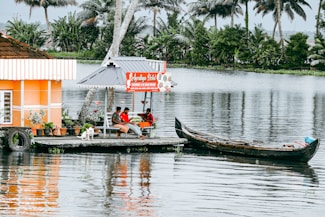 A peaceful riverside scene with a small dock where two people are seated under an open-air patio next to a sign that reads 'Ayodhya Hotel'. A dog is sitting nearby, and a traditional wooden boat is moored to the dock. Palm trees and lush greenery are visible across the calm water.