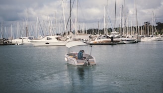 A small motorboat navigates through a marina filled with numerous sailboats and yachts docked at the piers. The water reflects the overcast sky, adding to the tranquil setting. A person is seated at the rear of the motorboat, steering it through the calm waters.