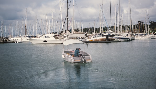 A small motorboat navigates through a marina filled with numerous sailboats and yachts docked at the piers. The water reflects the overcast sky, adding to the tranquil setting. A person is seated at the rear of the motorboat, steering it through the calm waters.