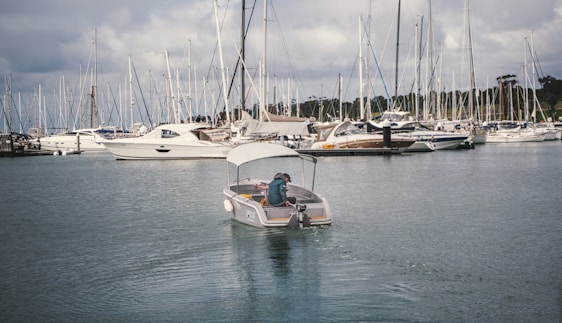 A small motorboat navigates through a marina filled with numerous sailboats and yachts docked at the piers. The water reflects the overcast sky, adding to the tranquil setting. A person is seated at the rear of the motorboat, steering it through the calm waters.