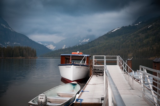 A tranquil mountain lake scene with a dock extending out into the water. Two boats are moored at the dock, one larger boat named Sinopah and a smaller rowboat with an outboard motor. The backdrop features dense forests and mist-covered mountains under a cloudy sky.