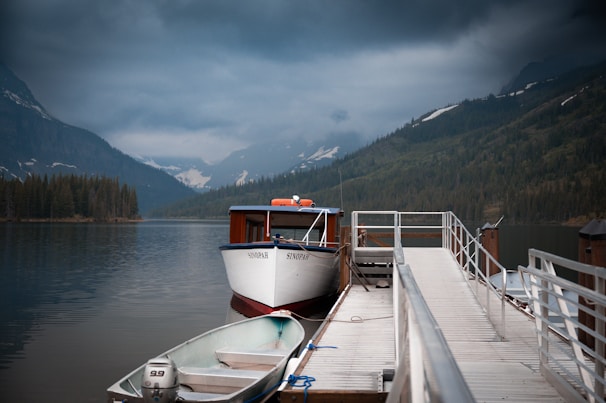 A tranquil mountain lake scene with a dock extending out into the water. Two boats are moored at the dock, one larger boat named Sinopah and a smaller rowboat with an outboard motor. The backdrop features dense forests and mist-covered mountains under a cloudy sky.