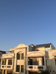 A residential building with a cream-colored facade is seen under a clear blue sky. The structure features multiple windows, balconies, and a rooftop area equipped with a staircase leading to an upper section. The sunlight casts warm shadows on the building, highlighting its architectural details.