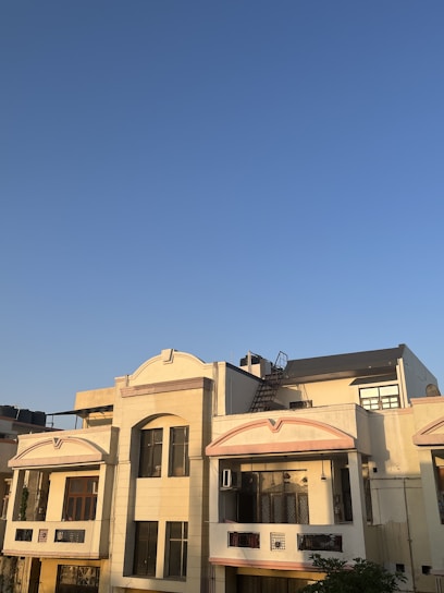 A residential building with a cream-colored facade is seen under a clear blue sky. The structure features multiple windows, balconies, and a rooftop area equipped with a staircase leading to an upper section. The sunlight casts warm shadows on the building, highlighting its architectural details.