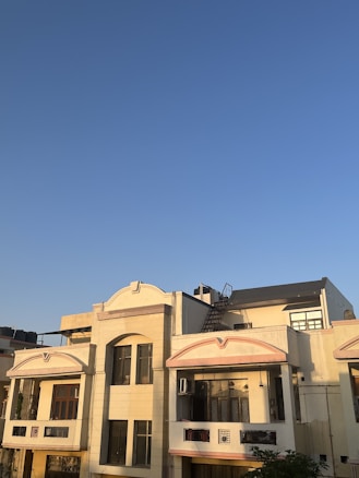 A residential building with a cream-colored facade is seen under a clear blue sky. The structure features multiple windows, balconies, and a rooftop area equipped with a staircase leading to an upper section. The sunlight casts warm shadows on the building, highlighting its architectural details.