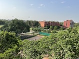 A lush, green landscape with dense trees in the foreground, surrounding a tennis court. In the background, brick buildings and a clear blue sky are visible, suggesting an educational or recreational facility. Tall floodlights tower over some of the trees, indicating sports facilities nearby.