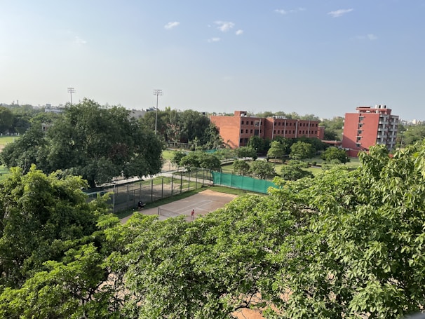 A lush, green landscape with dense trees in the foreground, surrounding a tennis court. In the background, brick buildings and a clear blue sky are visible, suggesting an educational or recreational facility. Tall floodlights tower over some of the trees, indicating sports facilities nearby.