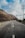A smooth asphalt road winds through a hilly landscape, flanked by utility poles and sparse vegetation. The sky is partly cloudy, casting shadows on the terrain. The road features a blue and yellow striped barricade on one side.