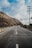 A smooth asphalt road winds through a hilly landscape, flanked by utility poles and sparse vegetation. The sky is partly cloudy, casting shadows on the terrain. The road features a blue and yellow striped barricade on one side.