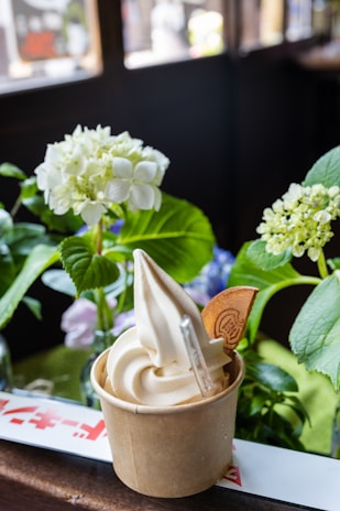 A cup of vanilla soft serve ice cream is placed on a wooden surface. The ice cream is swirled and decorated with a thin waffle biscuit. In the background, there are lush green leaves and white hydrangea flowers, adding a natural touch.