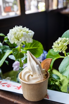 A cup of vanilla soft serve ice cream is placed on a wooden surface. The ice cream is swirled and decorated with a thin waffle biscuit. In the background, there are lush green leaves and white hydrangea flowers, adding a natural touch.