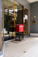 A modern glass storefront features a prominent red mailbox on a black stand placed on a welcome mat. The reflection of an old-style building with a tiled roof can be seen in the glass. Various signs and notices are displayed on the door and window. To the side, a small bunch of dried plants hangs on the wall.