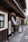 A traditional Japanese building with dark wooden panels and a white exterior. The entrance features a small sign with Japanese characters and a hanging fabric. A sidewalk runs alongside the building, leading to a sign with a portrait and an arrow directing visitors.