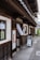 A traditional Japanese building with dark wooden panels and a white exterior. The entrance features a small sign with Japanese characters and a hanging fabric. A sidewalk runs alongside the building, leading to a sign with a portrait and an arrow directing visitors.