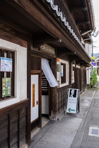 A traditional Japanese building with dark wooden panels and a white exterior. The entrance features a small sign with Japanese characters and a hanging fabric. A sidewalk runs alongside the building, leading to a sign with a portrait and an arrow directing visitors.