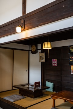 A traditional Japanese room features tatami flooring, a low wooden table, and cushions for seating. A vintage clock and framed artwork are mounted on the wall. The room is softly lit by a hanging lamp and a wall sconce.